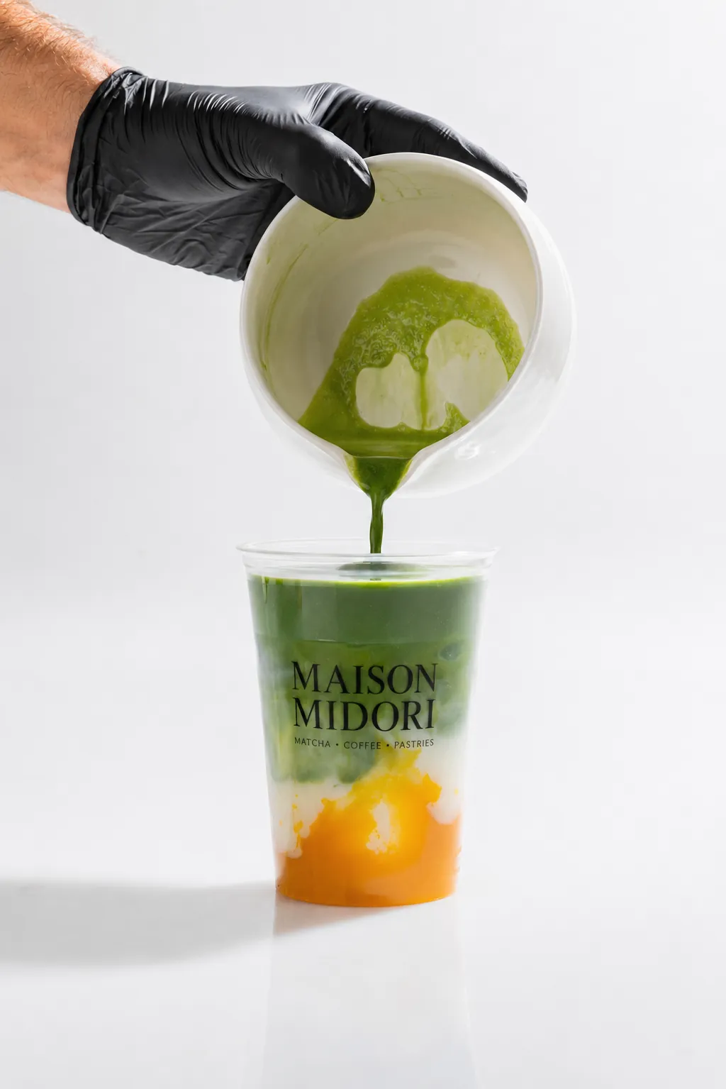 Barista mit schwarzem Handschuh gießt frisch gewhiskten Matcha aus einem Krug in einen Maison-Midori-Becher mit Mango-Schicht